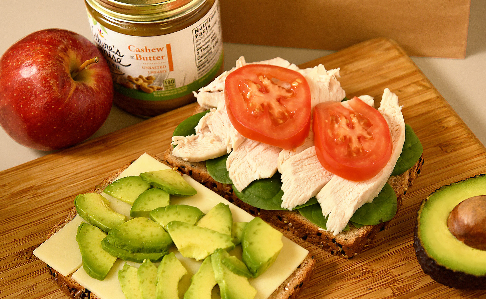 delicious sandwich, chicken breast, swiss cheese, sliced tomato, avocado and baby spinach on multi-grain wheat bread, also showing apple and cashew butter